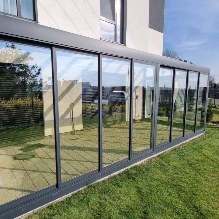 A modern, glass-enclosed aluminum conservatory attached to a single-family home with a green lawn.