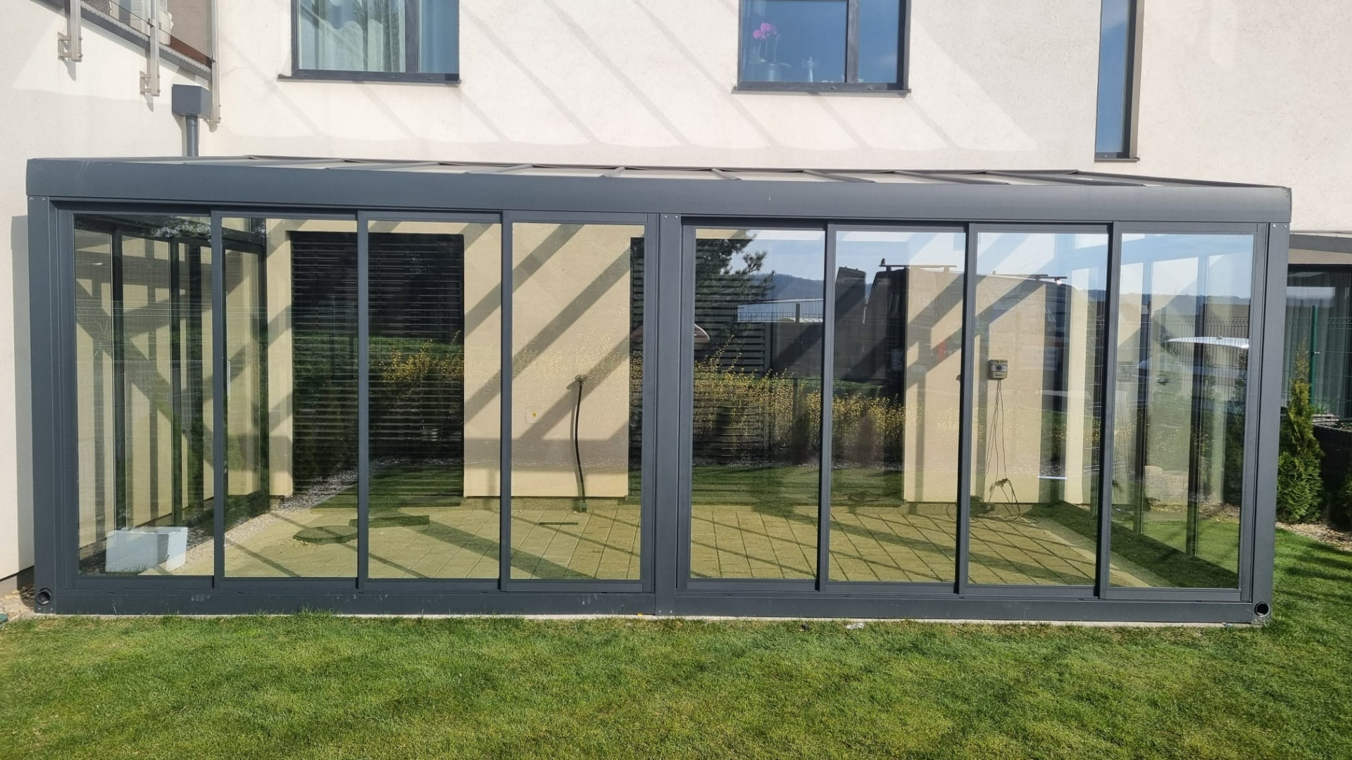 A modern, glass-enclosed aluminum sunroom attached to a single-family home with a view of the lawn.