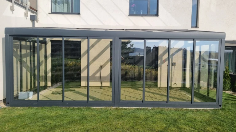 A modern, glass-enclosed aluminum sunroom attached to a single-family home with a view of the lawn.