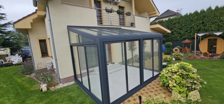 A modern, glass-enclosed sunroom with an anthracite-colored aluminum frame attached to a single-family home