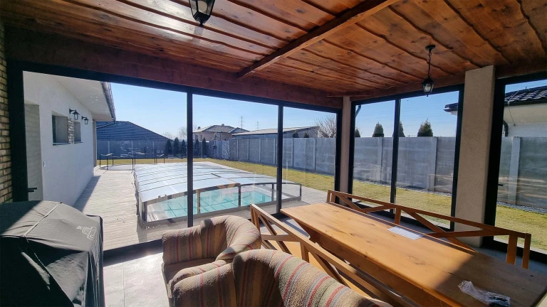 A view from inside the glass-enclosed terrace with a wooden ceiling out onto the garden with a pool