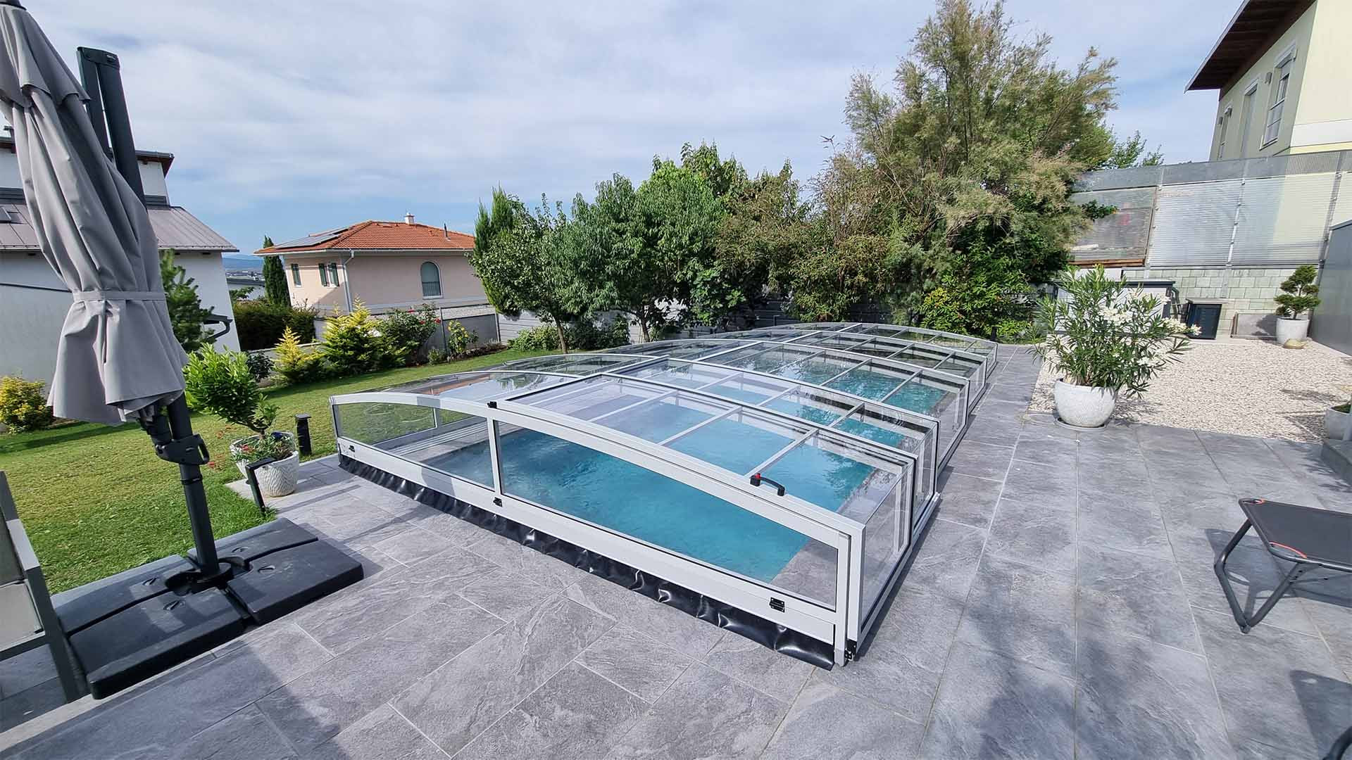 A modern, low-profile PREMIER aluminum pool enclosure on a paved patio in the garden of a single-family home