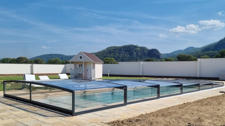 A modern, low-profile glass pool enclosure in the garden, with a view of the mountains and a garden shed.