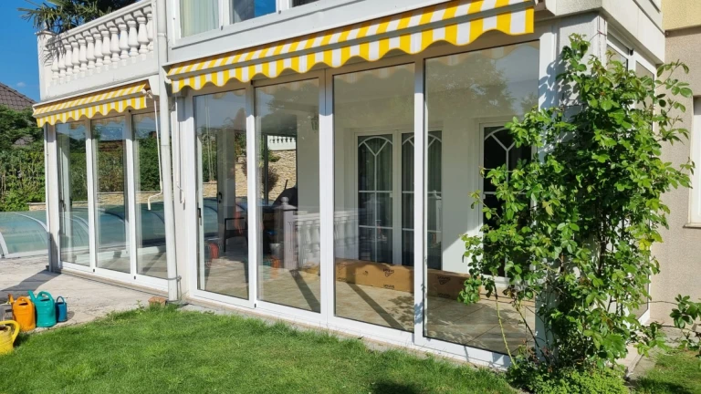 Modern terrace glazing with a white frame and a yellow-and-white awning in the sunny garden.