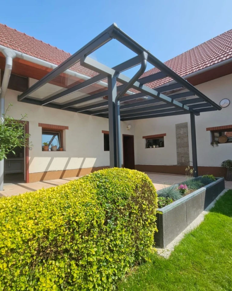 A modern aluminum pergola with a glass roof attached to a single-family home with a landscaped garden.