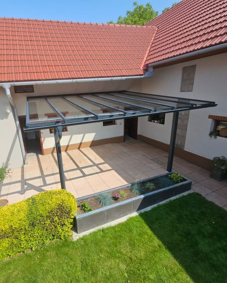 A modern aluminum pergola with a glass roof attached to the wall of the house, featuring a paved patio.