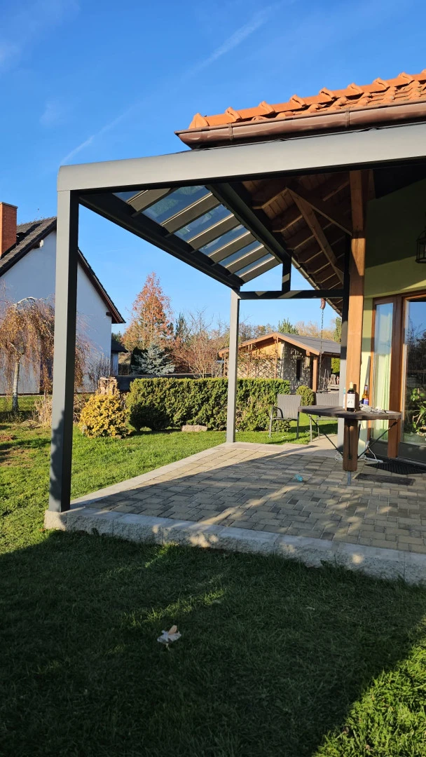 A modern gray aluminum pergola with a glass roof section on a paved patio next to a single-family home with a garden.