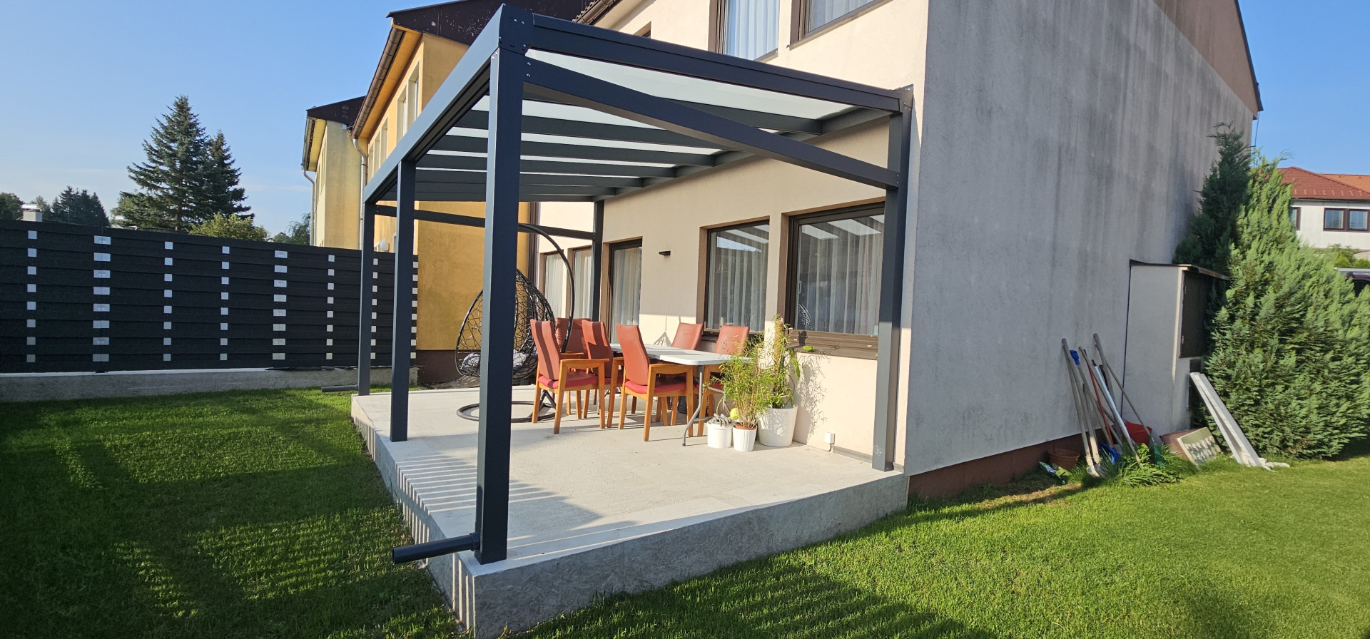 A modern aluminum pergola with a glass roof on the concrete patio of a single-family home, featuring garden furniture.