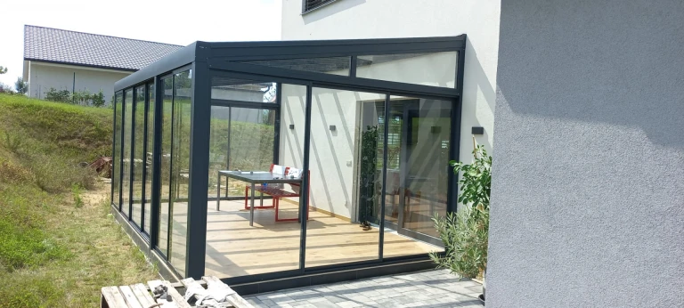 A modern glass-enclosed sunroom with an anthracite frame and sliding doors attached to a single-family home