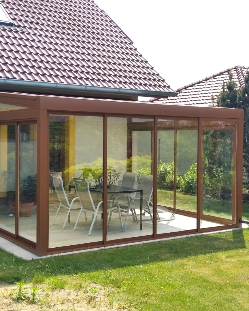 A glass-enclosed aluminum sunroom with a brown frame and patio furniture on the terrace next to the house