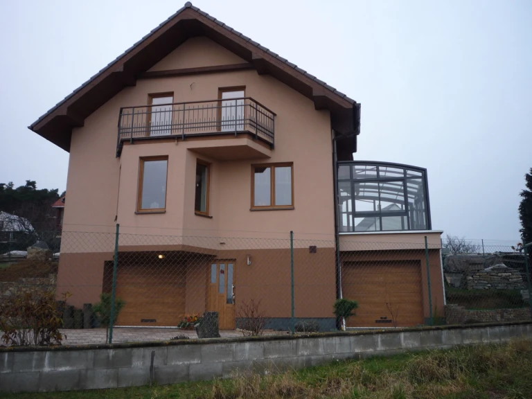 A view of a single-family home with a glass-enclosed sliding sunroom extension on the terrace above the garage.