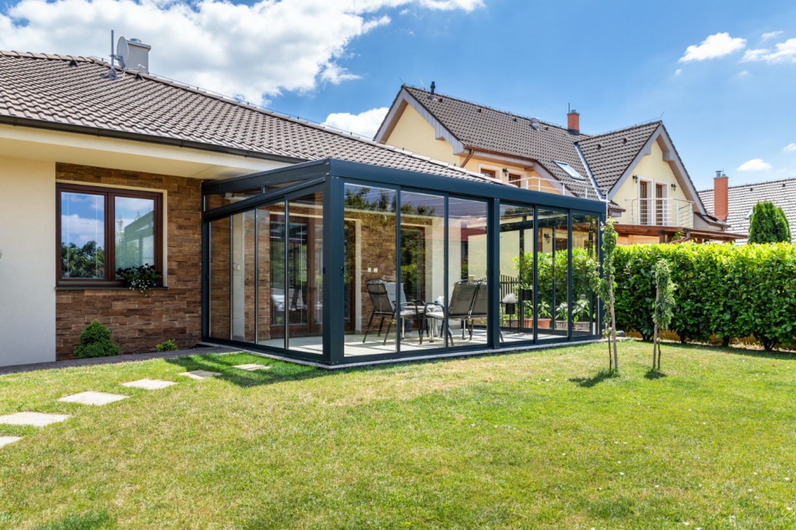 A modern, glass-enclosed sunroom with a dark frame, attached to a single-family home and furnished with garden furniture.