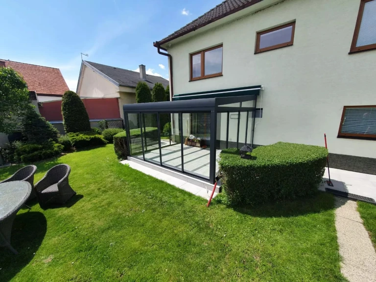 A modern, glass-enclosed sunroom with an anthracite-colored frame, attached to a single-family home with a garden.