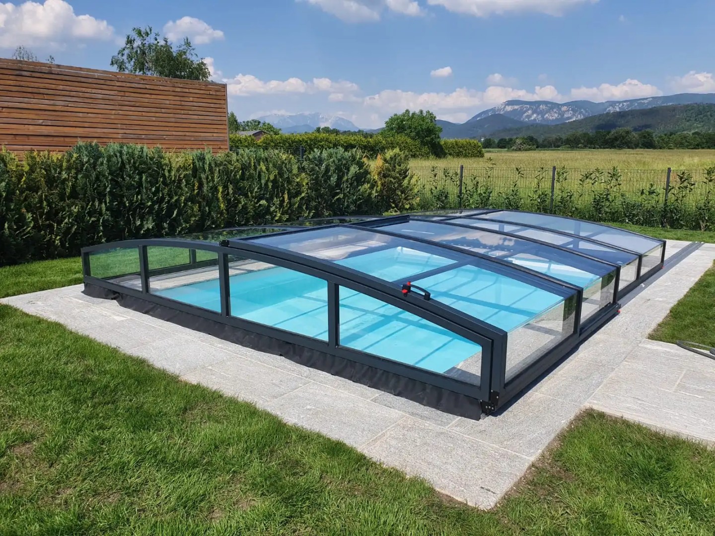 A low, anthracite-colored STAR pool enclosure in a garden with a view of the mountains