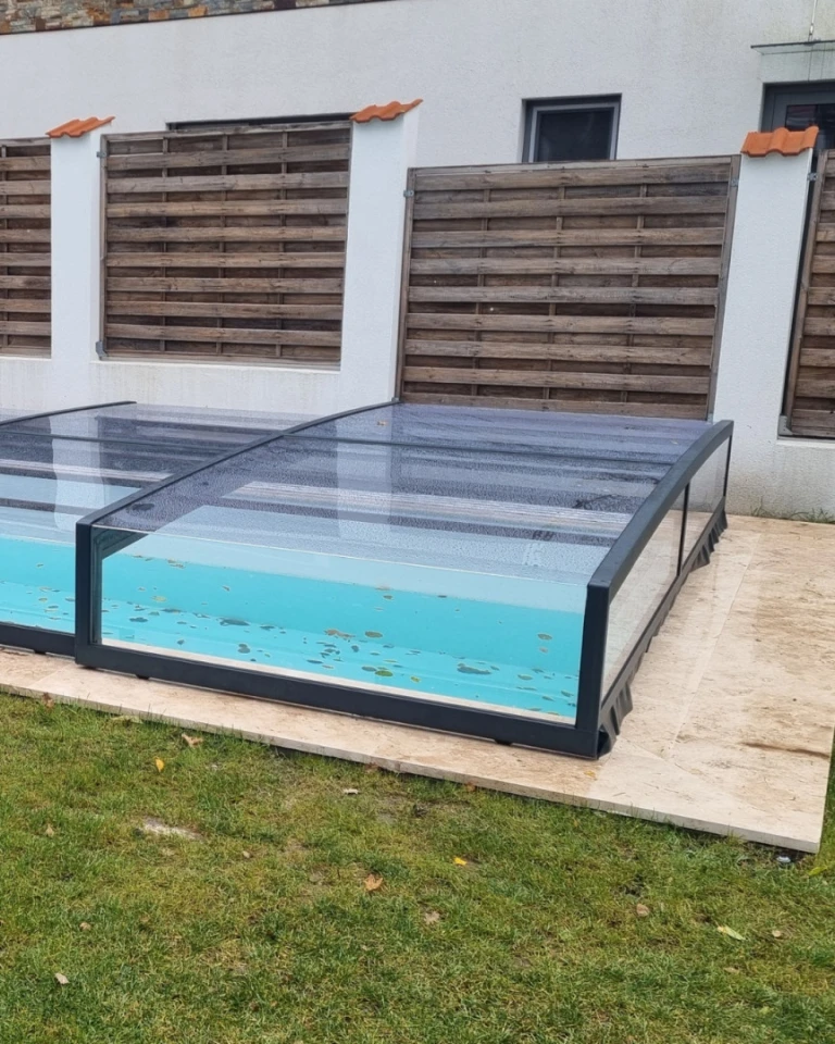 A low, anthracite-colored pool enclosure with clear polycarbonate panels next to the house