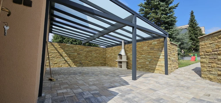 A modern aluminum patio roof with a glass-enclosed central section, stone-clad walls, and an outdoor fireplace.