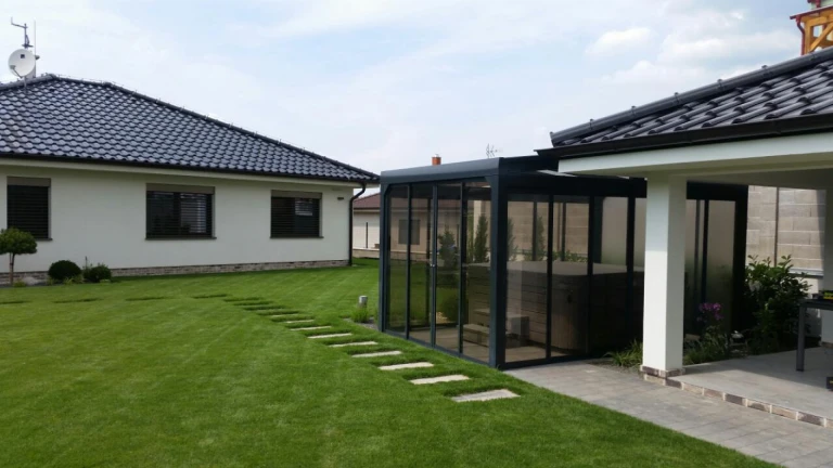 A modern glass-enclosed pavilion with a hot tub in a landscaped garden next to a single-family home.
