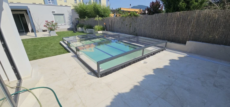 A modern, low-profile EXCLUSIVE pool enclosure with a dark frame and clear panels on the patio of a single-family home