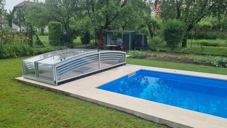 A modern, low-profile pool enclosure with a transparent panel next to an open-air pool in the garden.