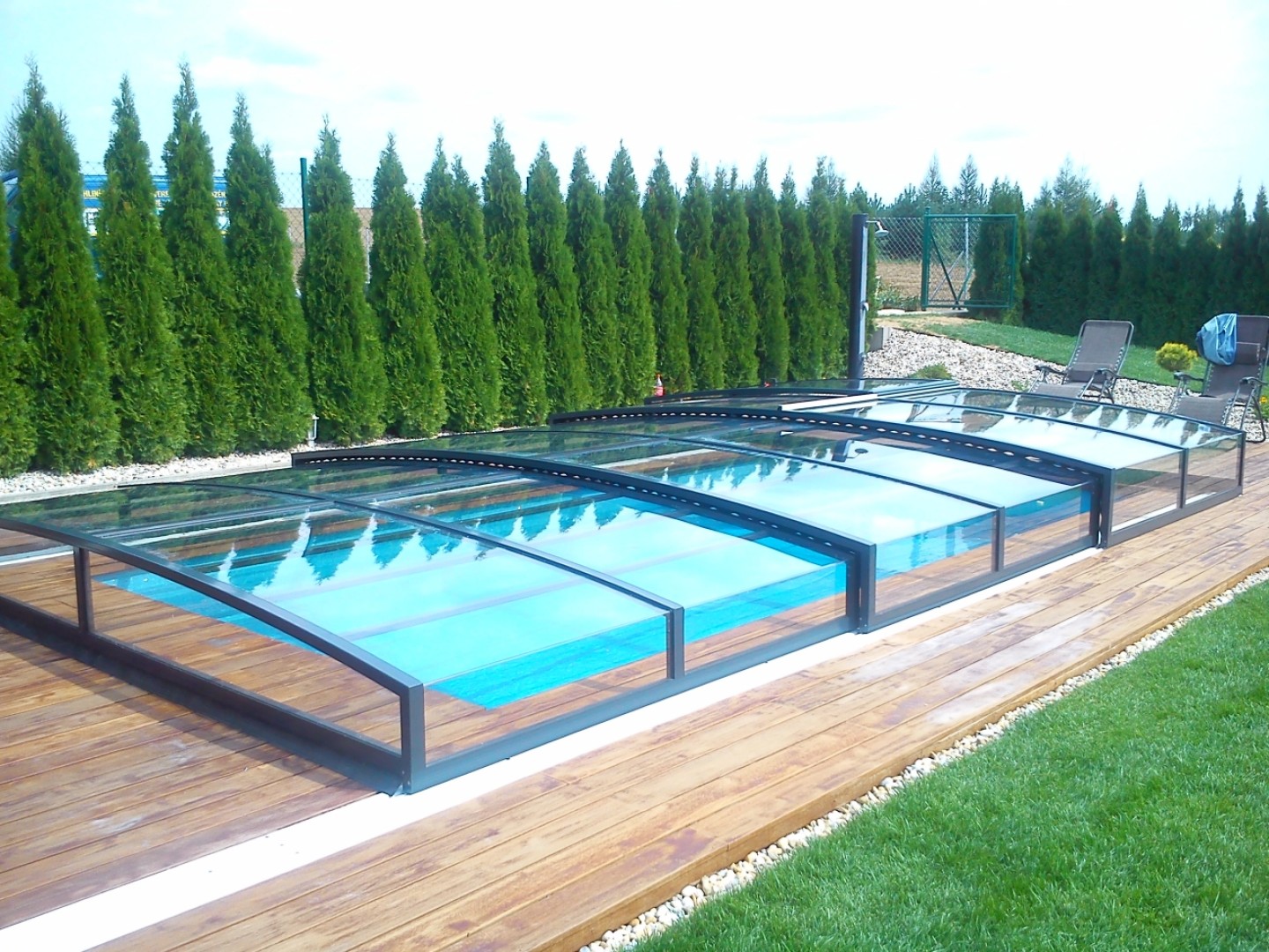 A modern, low-profile pool enclosure with a transparent panel on a wooden deck surrounded by greenery