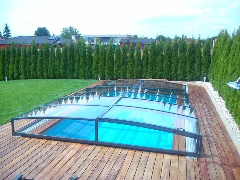 A modern, low-profile pool enclosure with a clear panel on a wooden deck in a garden with a hedge.