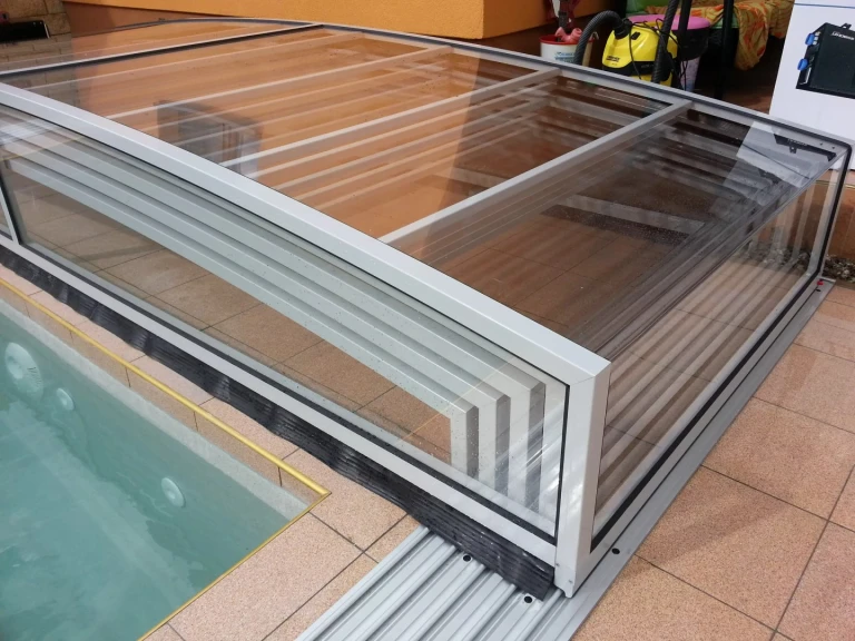 A modern, low-profile pool enclosure with an aluminum frame and a transparent covering over the pool.