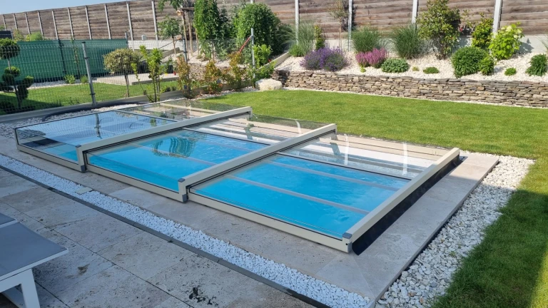 A modern, low-profile telescopic pool enclosure with glass panels in a landscaped garden.