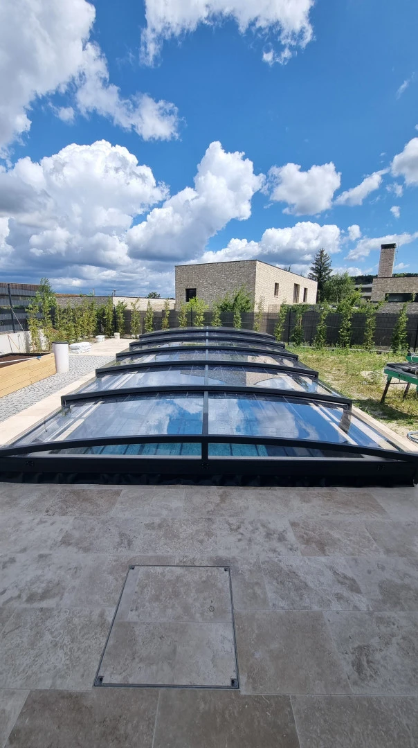 A modern, low-profile pool enclosure with a black frame and transparent panels in the garden of a single-family home.