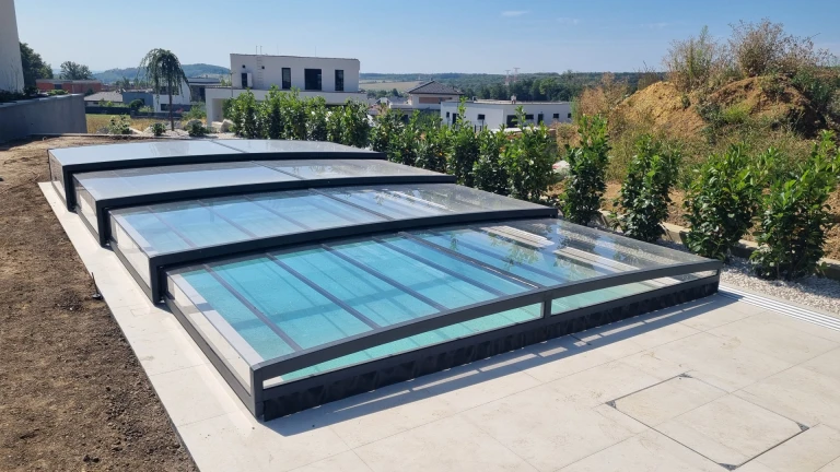 A modern, low-profile telescopic pool enclosure with an anthracite frame and transparent panels on the terrace of a single-family home.