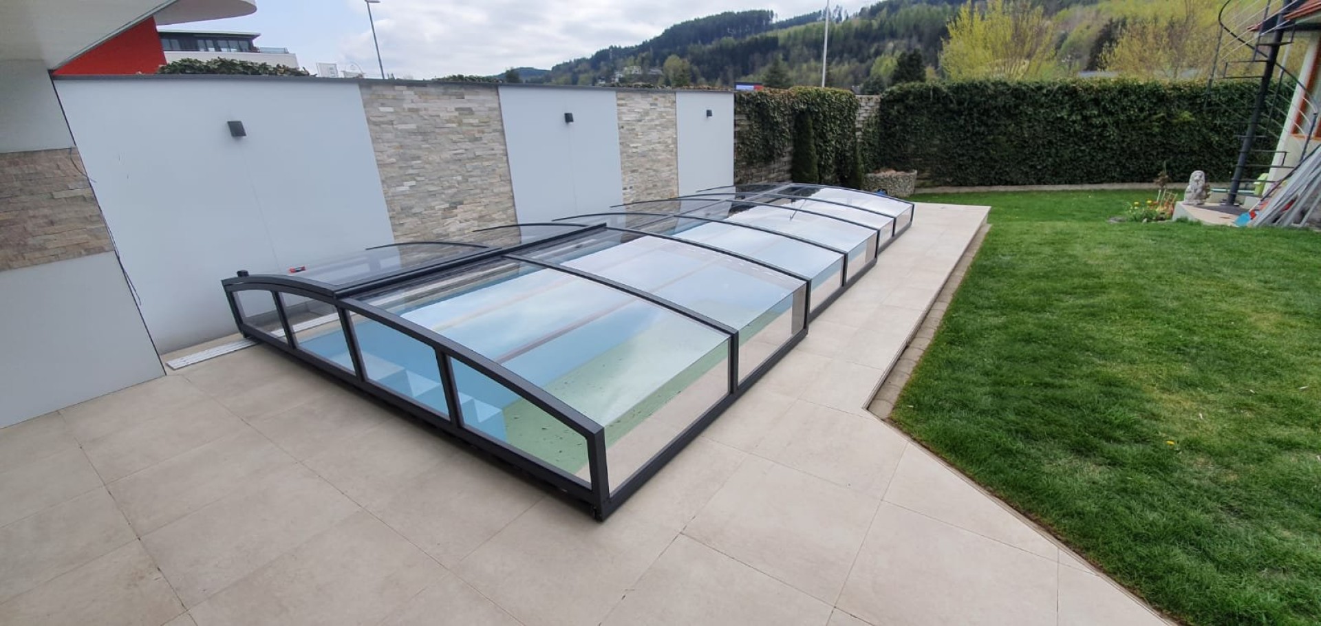 A modern, low-profile pool enclosure with an anthracite frame on the terrace next to the garden