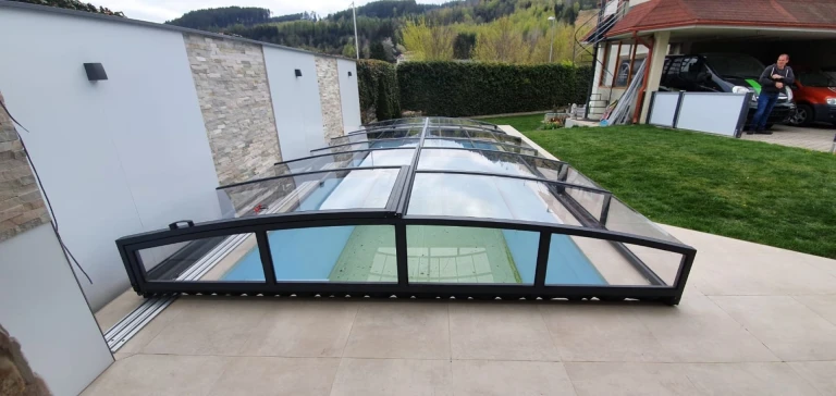 A modern, low-profile pool enclosure with a black frame and glass panels in the garden