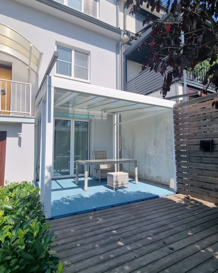 A white, modern aluminum pergola with a glass roof on the patio of a single-family home, featuring garden furniture.