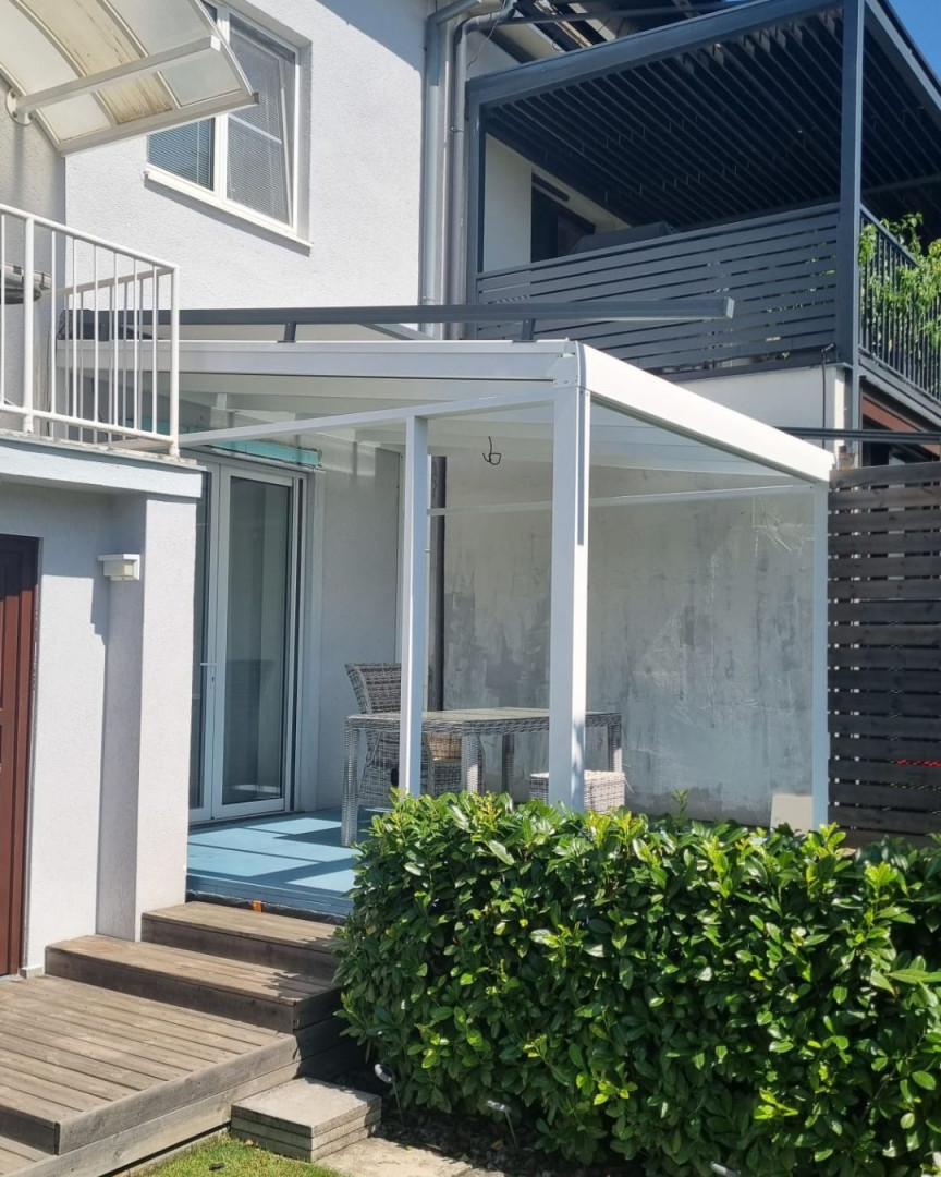 A white, modern aluminum pergola with a glass roof next to a single-family home, featuring patio furniture.
