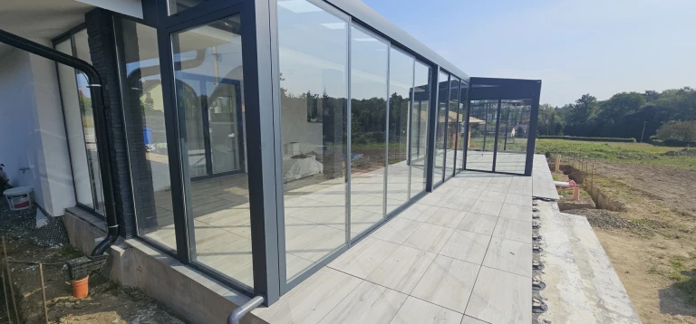 A modern glass-enclosed sunroom with an anthracite aluminum frame and a paved terrace.