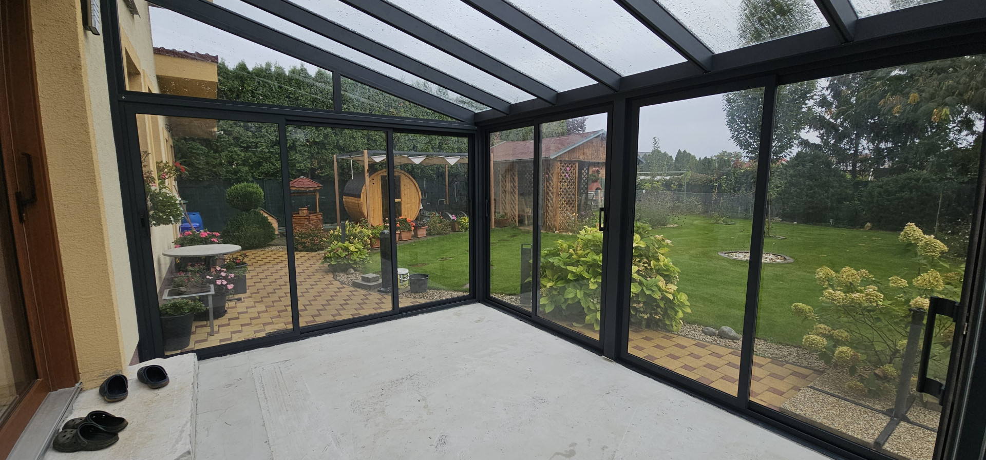 A modern, glass-enclosed sunroom with a black aluminum frame and a view of the backyard, which features a sauna