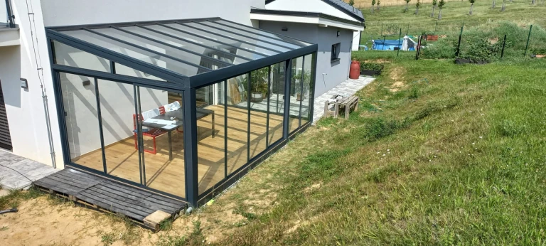 A modern glass-enclosed sunroom with an anthracite-colored frame and wooden flooring attached to a single-family home