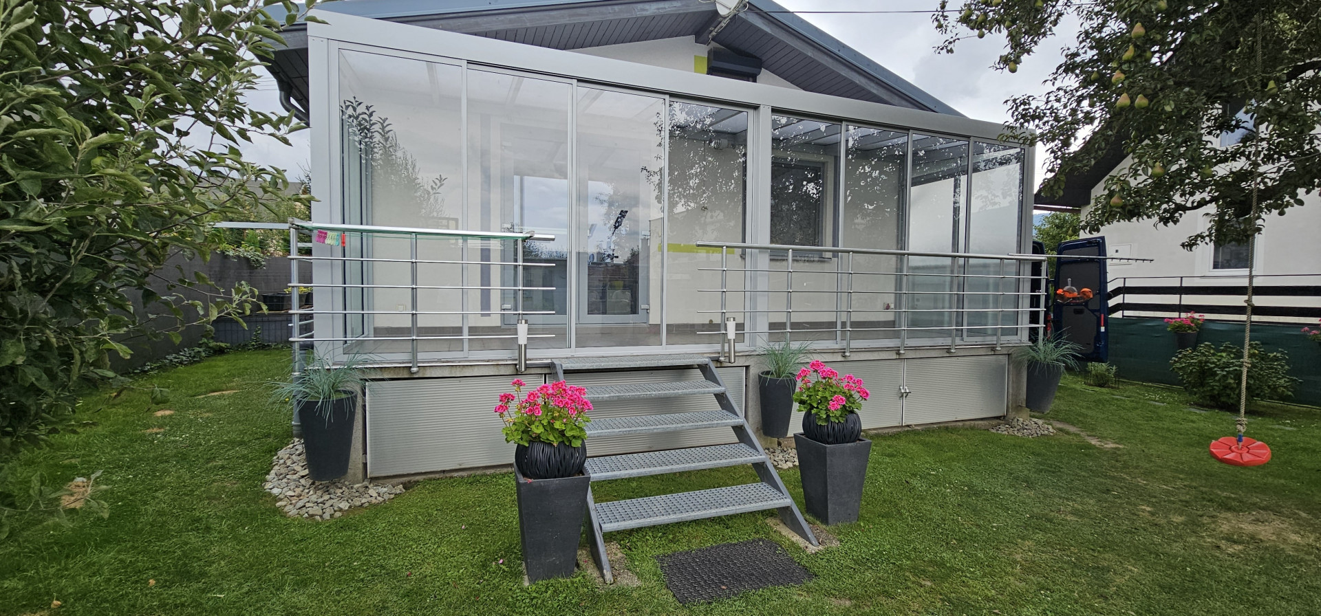 A modern, glass-enclosed sunroom on a raised terrace with stairs and floral decorations