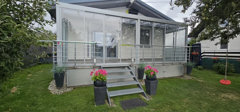A modern, glass-enclosed sunroom on a raised terrace with stairs and floral decorations