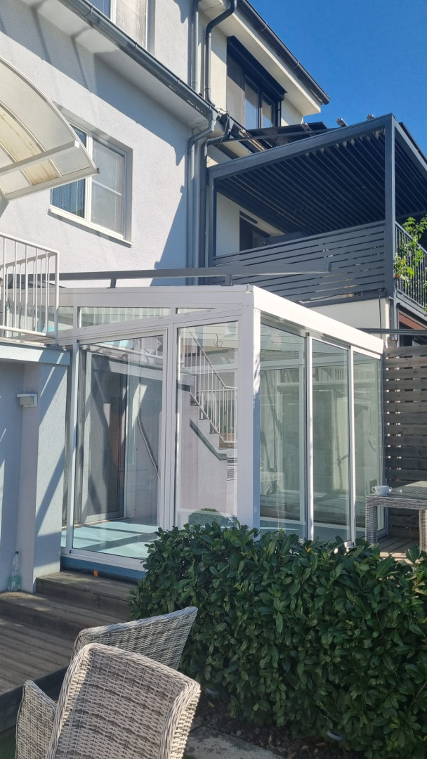 A modern white glass-enclosed sunroom attached to a single-family home, with a view of the patio and the greenery.
