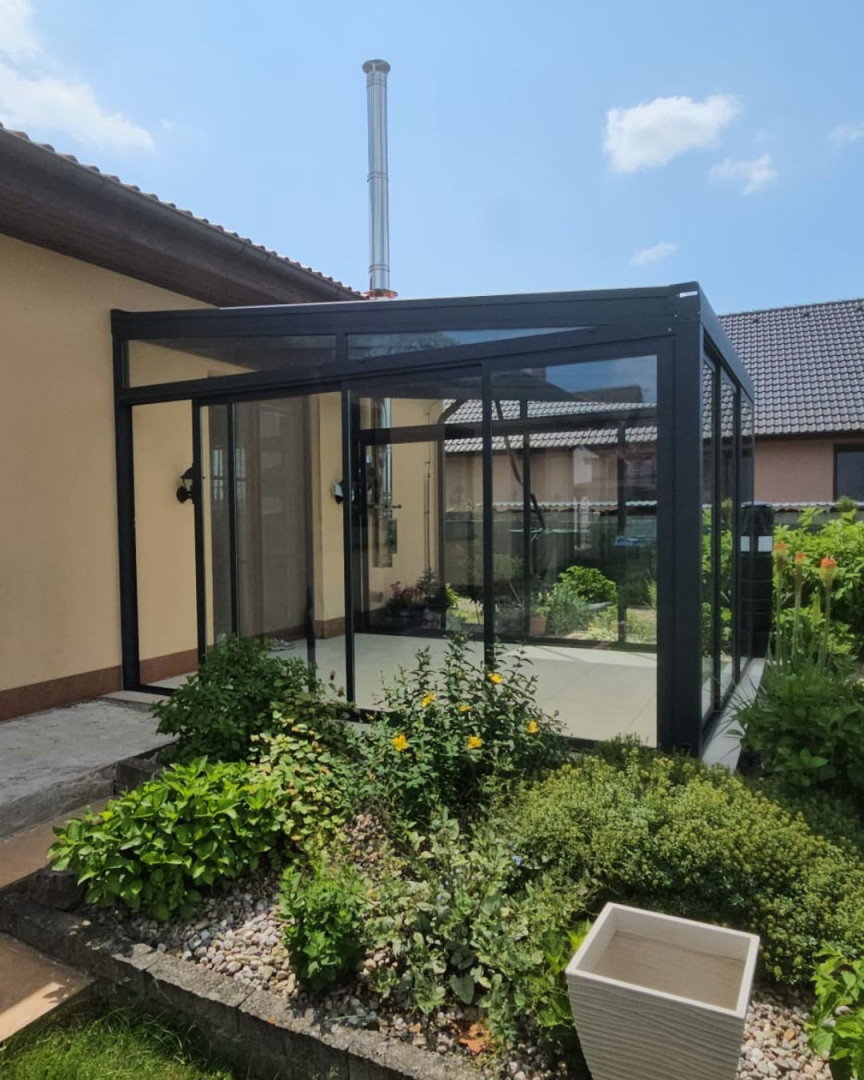 A modern glass addition to a single-family home with black frames and sliding doors, surrounded by a garden.