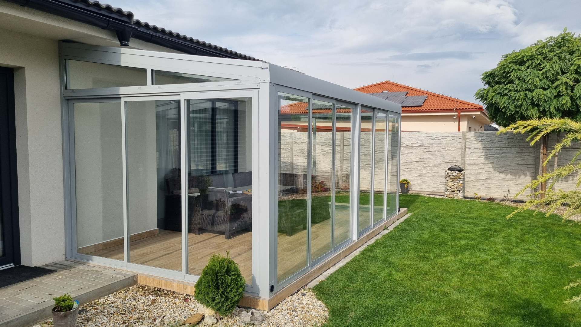 A modern, glass-enclosed sunroom with an aluminum frame attached to a single-family home with a garden
