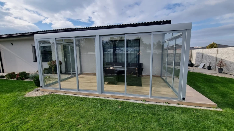 A modern, glass-enclosed sunroom with a gray frame on a wooden deck next to a single-family home