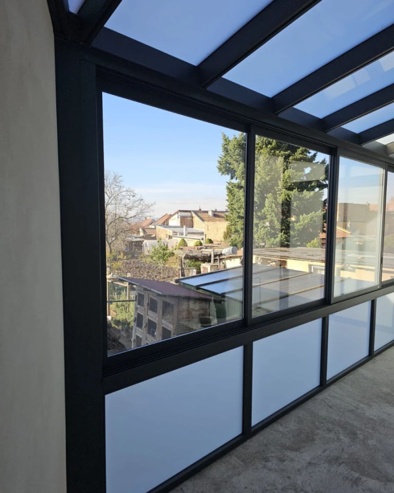 A view through the glass wall of the black-framed sunroom of the surrounding buildings and gardens.