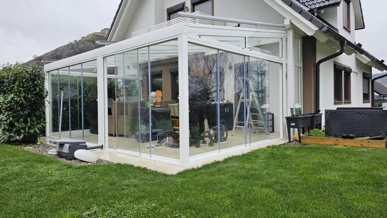 A modern, glass-enclosed white aluminum sunroom attached to a single-family home with a garden