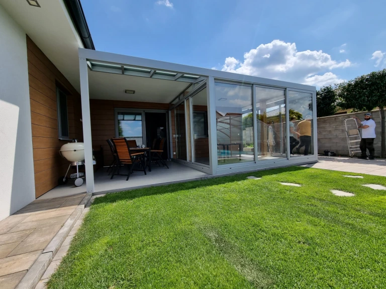 A modern, glass-enclosed sunroom with an aluminum frame, built next to a single-family home with a well-manicured lawn.