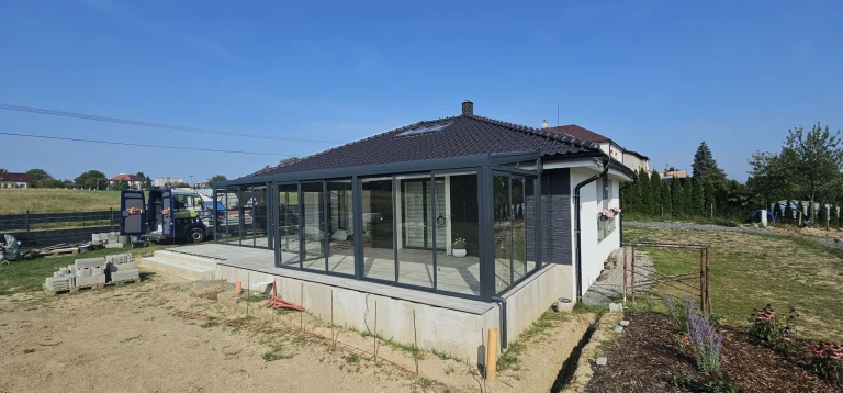 A modern, glass-enclosed sunroom with an anthracite-colored frame, built next to a single-family home