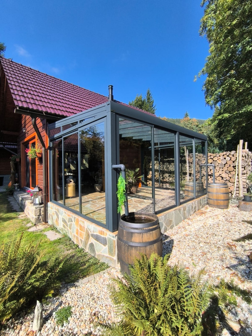 A modern, glass-enclosed sunroom with a dark aluminum frame, attached to a wooden house with a stone foundation.