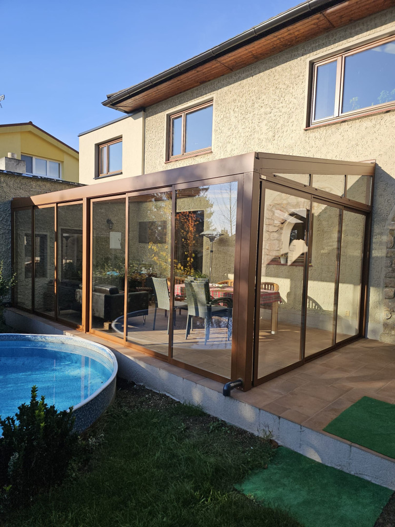 A modern, glass-enclosed sunroom with a brown frame attached to a single-family home with a pool