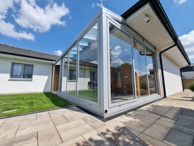 A modern, glass-enclosed sunroom with an aluminum frame attached to a single-family home overlooking the lawn