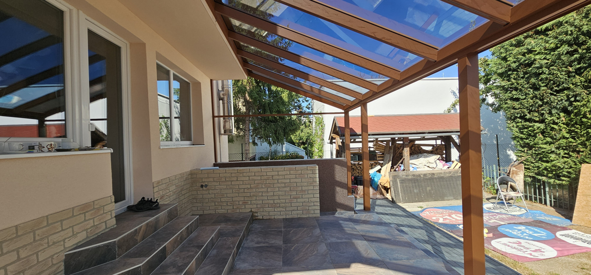 An aluminum pergola with a wood-grain finish and a glass roof, installed on the deck of a single-family home.